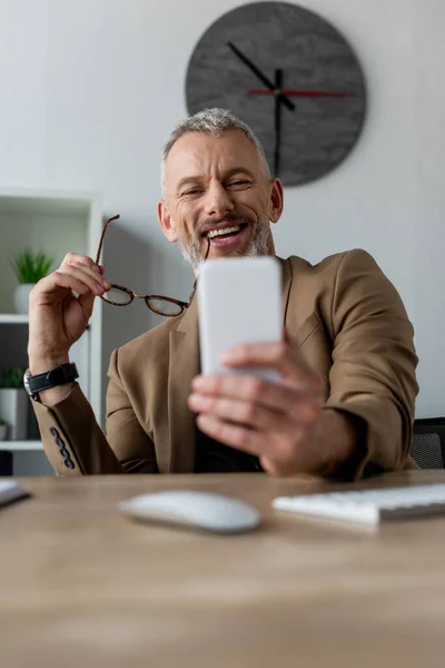 Enfoque selectivo de empresario feliz sosteniendo gafas y mirando el teléfono inteligente en la oficina - foto de stock