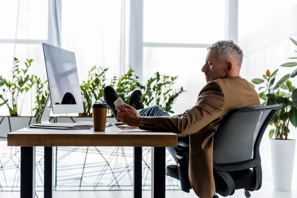 Вид сбоку красивого бизнесмена, сидящего у монитора компьютера и держащего смартфон — стоковое фото