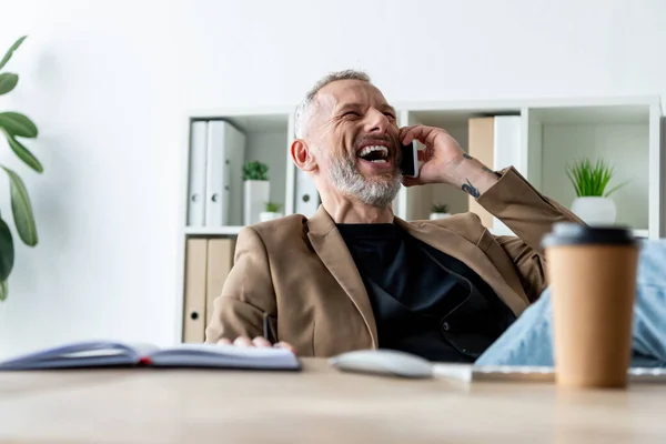 Enfoque selectivo de hombre de negocios alegre hablando en el teléfono inteligente cerca de la taza de papel - foto de stock