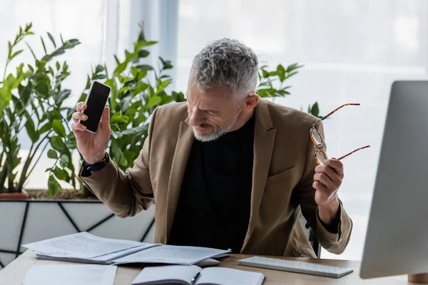 Bearded businessman holding glasses and smartphone with blank screen while looking at documents near computer monitor — Stock Photo