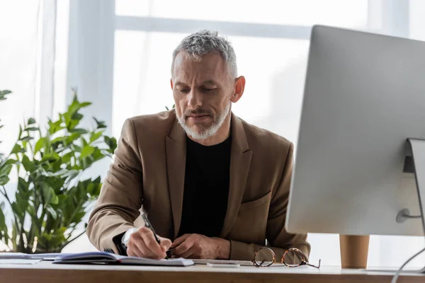 Bearded businessman writing in notebook near computer monitor — Stock Photo