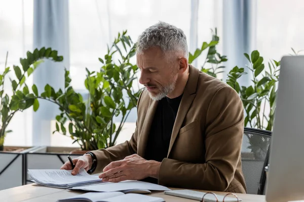 Bearded businessman looking at document while sitting at desk in office — Stock Photo