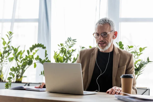 Bel homme d'affaires en lunettes et écouteurs regardant ordinateur portable près de tasse en papier sur la table — Photo de stock