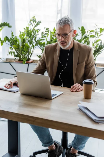 Bearded businessman in glasses and earphones looking at laptop near paper cup on table — Stock Photo