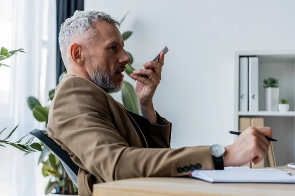 Vue latérale d'un homme d'affaires barbu enregistrant un message vocal tout en tenant un smartphone — Photo de stock