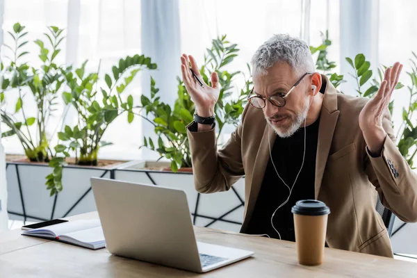 Confuso uomo d'affari in occhiali e auricolari che mostra gesto scrollata di spalle e guardando il computer portatile mentre ha videochiamata — Foto stock