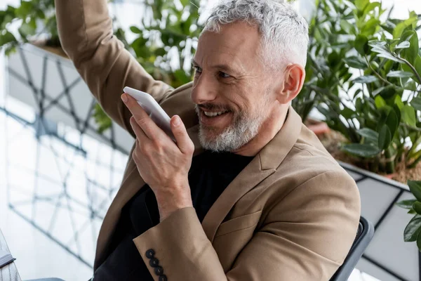 Cheerful businessman recording voice message near green plants in office — Stock Photo