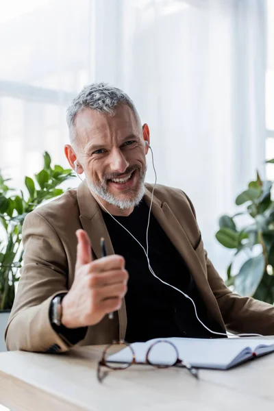 Selective focus of happy businessman in earphones showing thumb up in office — Stock Photo