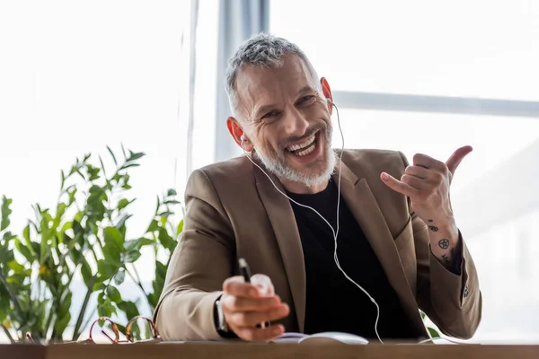 Selective focus of happy businessman in earphones looking at camera and showing call me gesture — Stock Photo