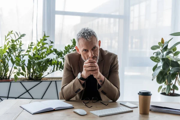 Homme d'affaires avec les mains serrées regardant la caméra près du smartphone et tasse de papier dans le bureau — Photo de stock