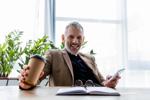 Selective focus of cheerful businessman holding paper cup and smartphone — Stock Photo