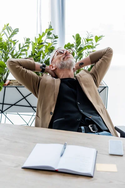 Selektiver Fokus des Geschäftsmannes in Brille, der nach oben schaut, während er neben Smartphone mit leerem Bildschirm und Notebook auf dem Tisch sitzt — Stockfoto