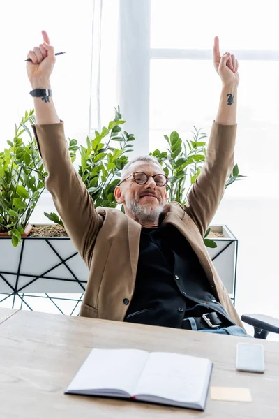 Selektiver Fokus glücklicher Geschäftsleute mit Händen über dem Kopf, die mit Fingern in der Nähe von Smartphones mit leerem Bildschirm und Notizbuch auf dem Tisch zeigen — Stockfoto