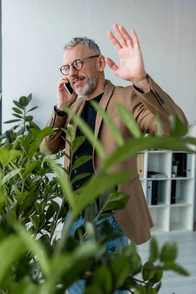 Selective focus of happy businessman waving hand while talking on smartphone near plants — Stock Photo