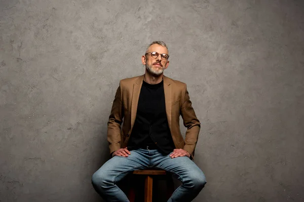 Bärtiger Geschäftsmann mit Brille blickt in die Kamera und sitzt auf grau — Stockfoto