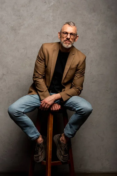 Hombre de negocios barbudo en gafas mirando a la cámara y sentado en el taburete en gris - foto de stock