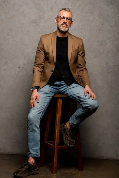 Businessman in glasses sitting on stool and looking at camera on grey — Stock Photo