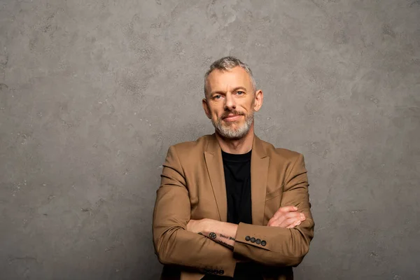 Bearded businessman standing with crossed arms and looking at camera on grey — Stock Photo