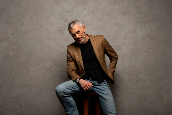 Empresário em jeans e blazer sentado no banquinho e olhando para a câmera em cinza — Fotografia de Stock
