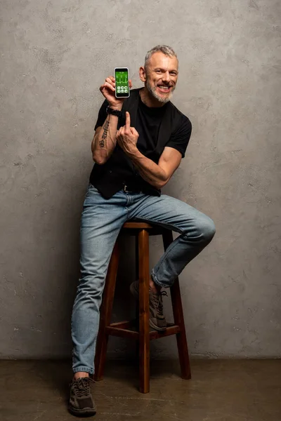 Uomo felice con tatuaggio puntato con il dito verso smartphone con app medica su grigio — Foto stock