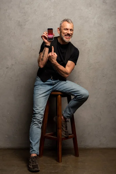 Hombre feliz con tatuaje apuntando con el dedo en el teléfono inteligente con cursos de comercio en gris - foto de stock