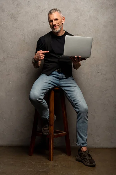 Счастливый мужчина показывает пальцем на ноутбук и смотрит на камеру на сером — стоковое фото