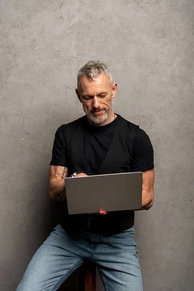 Hombre guapo con tatuaje usando el ordenador portátil en gris - foto de stock