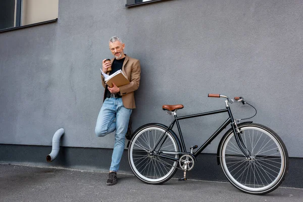 Bel homme d'affaires en blazer et jeans debout près du vélo tout en tenant la tasse en papier et le dossier — Photo de stock
