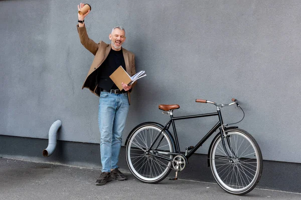 Homem de negócios feliz acenando mão enquanto segurando copo de papel e pasta e de pé perto de bicicleta — Fotografia de Stock