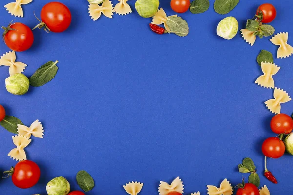 Colorful pizza ingredients pattern frame made of cherry tomatoes, basil and farfalle on blue background. Cooking concept.