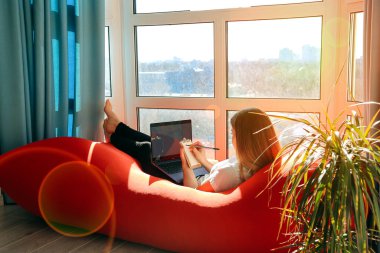 Evde çalış. Dizüstü bilgisayarda çalışan güzel, genç bir kadın pencerenin yanındaki büyük, rahat sandalyede otururken.