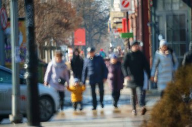 Arka plan bulanık. Çocuğu olan bir anne şehir caddesi boyunca yürür. Şehir hayatı. Kalabalığın hareketi. İnsan siluetleri. Aile tatili. 