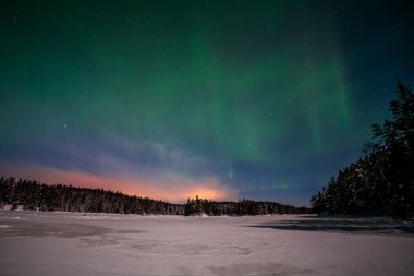 Dramatik kutup ışıkları, İsveç 'te donmuş bir göl ve karlı bir ormanın üzerinde ay ışığında gökyüzünde birçok bulut ve yıldızla Aurora borealis. uzun deklanşör hızı.