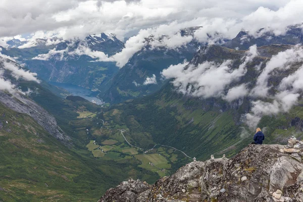 Een eenzaam jong meisje zit aan de rand van een berg en geniet van het uitzicht op de Geiranger fjord en adelaarsweg bij bewolkt weer van Dalsnibba berg, serpentine weg, Noorwegen, selectieve focus. — Stockfoto