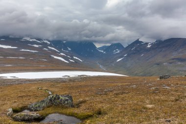 İsveç Laponya 'daki Sarek ulusal parkının güzel vahşi doğası. Karlı dağ zirveleri, nehir ve göl, huş ağacı ve çam ağaçları ormanı. Fırtınalı havada sonbahar renkleri. seçici odak