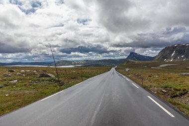 Bulutlu gökyüzünün altında Norveç dağları arasında ıslak düz ve ıssız bir yol