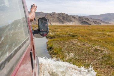 Turistin eli arabanın camından dışarı sarkmış, manzaranın fotoğrafını çekmiş. SUV arabasıyla nehir yolculuğu. Moğolistan - Altai. Fotoğraf içeriden çekilmiş.