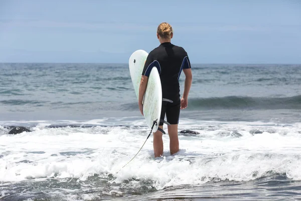 O surf entra na água. Homem surfista entrando no mar com sua prancha em um terno de surf preto. Tenerife, Espanha . — Fotografia de Stock
