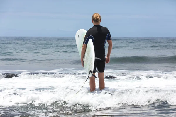 Die Brandung dringt ins Wasser ein. Männlicher Surfer, der mit seinem Brett in einem schwarzen Surfanzug aufs Meer steigt. Teneriffa, Spanien. lizenzfreie Stockbilder