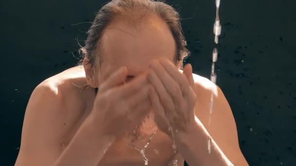 Un hombre caucásico, desnudo hasta la cintura, se lava la cara con un chorro de agua. Fondo negro, cámara lenta . — Vídeos de Stock