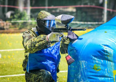 Koruyucu üniformalı ve maskeli bir paintball oyuncusu dışarıda silahla oynuyor.