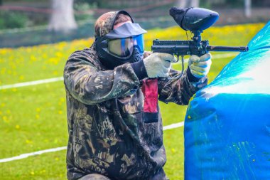 Koruyucu üniformalı ve maskeli bir paintball oyuncusu dışarıda silahla oynuyor.