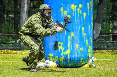 Koruyucu üniformalı ve maskeli bir paintball oyuncusu dışarıda silahla oynuyor.