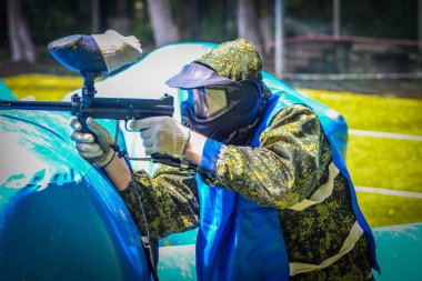 Koruyucu üniformalı ve maskeli bir paintball oyuncusu dışarıda silahla oynuyor.