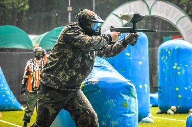 Koruyucu üniformalı ve maskeli bir paintball oyuncusu dışarıda silahla oynuyor.