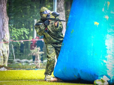 Koruyucu üniformalı ve maskeli bir paintball oyuncusu dışarıda silahla oynuyor.