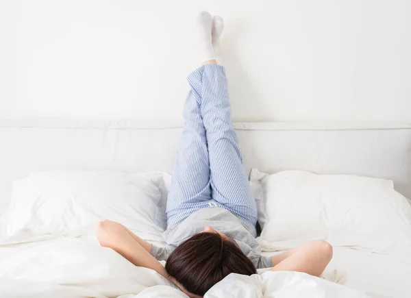 Woman with legs raised up high and arms under her head lying on bed in bedroom