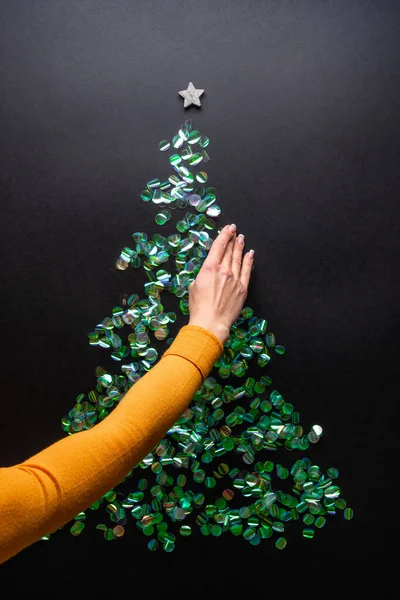 Mão feminina decorando árvore de Natal feita de confete verde no fundo preto. Conceito de ano novo com ouropel. Deitado plano, vista superior — Fotografia de Stock