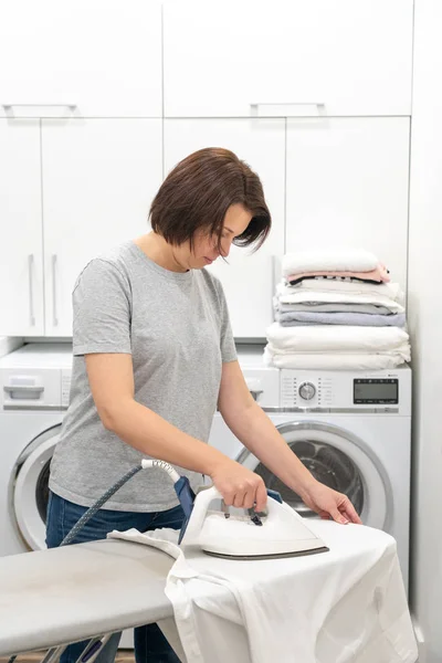 Woman ironing white shirt on board in laundry room with washing machine on background — Stock Photo, Image