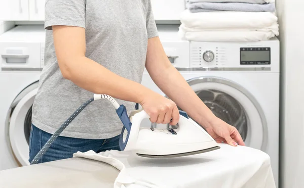 Жінка прасує білу сорочку на борту в пральній кімнаті з пральною машиною на фоні — стокове фото